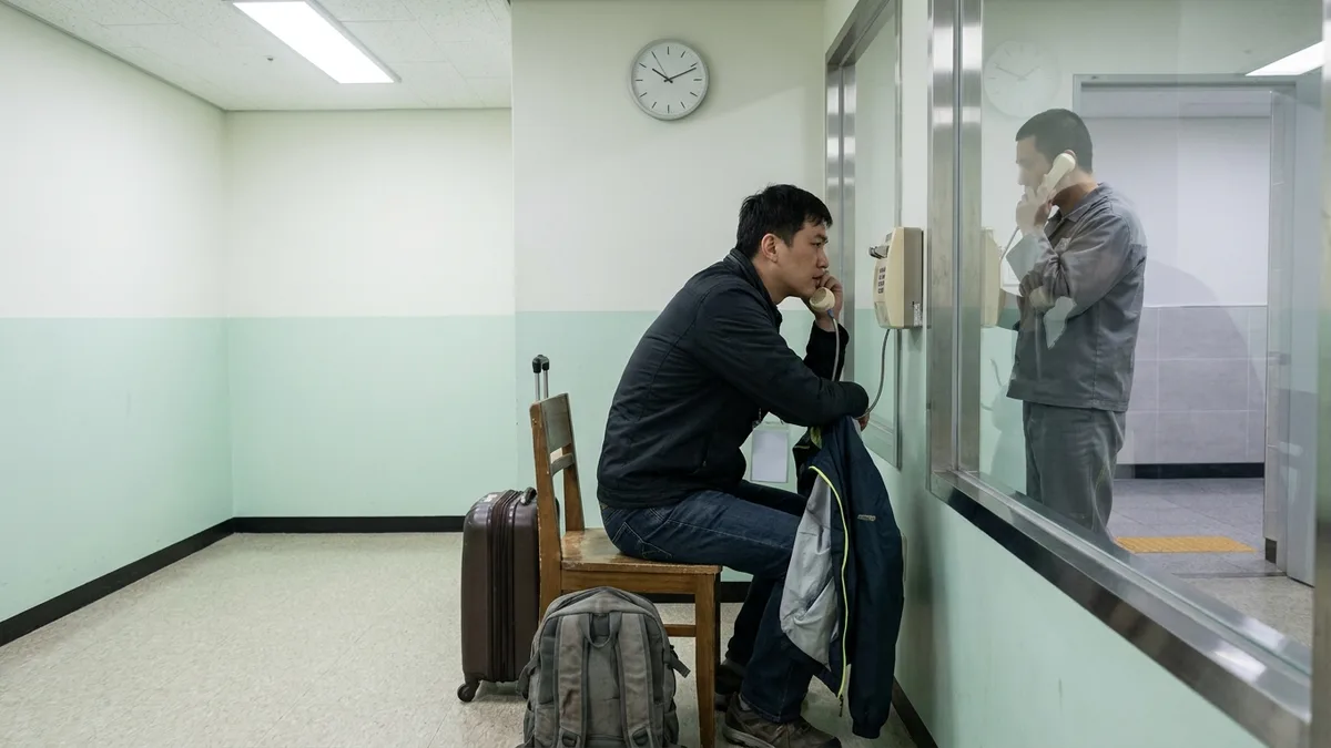 ภาพห้องพบปะในศูนย์พักพิง มีกระจกกั้นตรงกลาง ผู้มาเยี่ยมนั่งบนเก้าอี้พร้อมกระเป้าสัมภาระและกระเป้าเป้ข้างตัว ฝั่งตรงข้ามเป็นผู้ถูกพักพิงที่สวมเครื่องแบบสีเทายืนอยู่ ทั้งสองฝ่ายถือหูฟังติดผนังคุยกันอยู่ ถ่ายภาพจากด้านข้าง